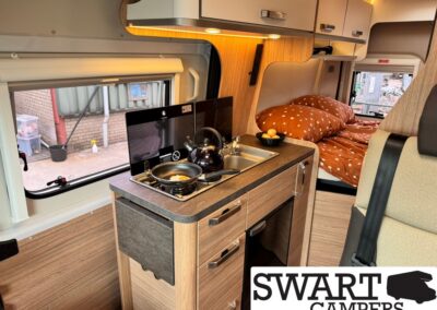 Cozy camper kitchenette with stove, kettle, sink, and wooden cabinetry, bed area visible in background, Swart Campers branding in corner.