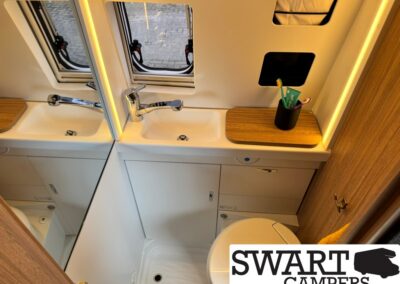 Compact camper bathroom with a white sink, chrome faucet, and a mirrored cabinet; wooden countertop with a toothbrush cup and toothpaste beside a small window.",