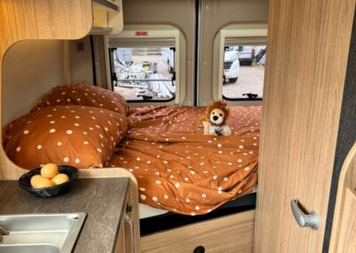 Compact camper van bedroom with a burnt‑orange polka dot duvet on a built‑in bed, a small lion plush toy, and a kitchenette counter with a double sink to the left; overhead cupboards and a skylight overhead, two rear windows show a parking area; SWART CAMPERS logo in the bottom right corner.