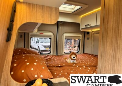 Cozy camper bedroom with brown polka-dot bedspread, a plush lion toy, and wooden cabinetry; skylight above and windows showing outside activity; Swart Campers branding in corner.