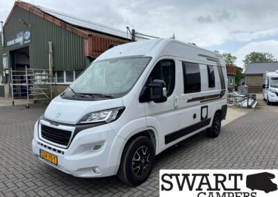 Front three-quarter view of a white camper van with black trim, parked on brick paving beside an industrial building and a SWART CAMPERS logo in the corner.