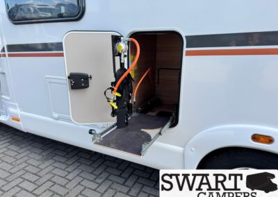 Wheelchair lift platform extended from the side of a white motorhome with an open door and orange hydraulic hoses visible inside the storage area