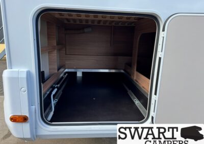 Open rear storage compartment of a camper van with wood-paneled interior, shelving on the left, and a black textured floor.