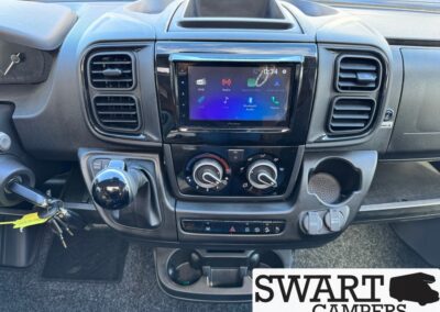 Dashboard of a camper van with a central touchscreen, round climate knobs, gear shifter on the left, and cupholders.