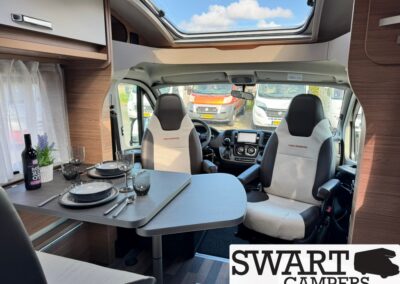 Camper van interior with a dining table set for two, wine bottle and glassware; front seats and dashboard visible under a skylight.
