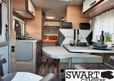 Interior of a modern camper van: compact kitchen left, dining table with seating, and a bed area visible in the background; wood cabinetry throughout.