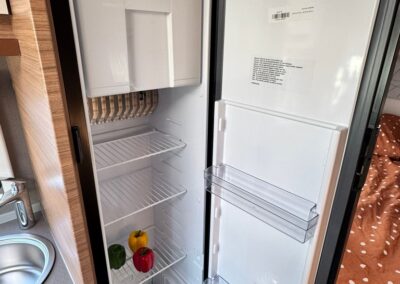 Open compact camper fridge with peppers on the lower rack and a can on the door shelf; sink and wood cabinetry nearby.