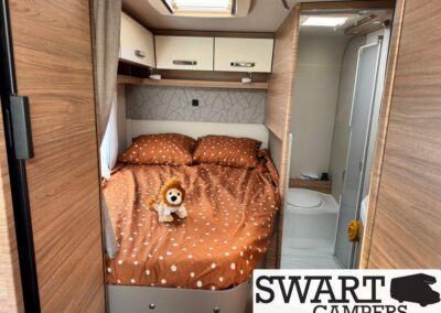 Camper bedroom with orange polka-dot bedding, a plush lion on the bed, and overhead cabinets; bathroom visible to the right.