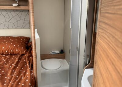 Compact camper bathroom with toilet, shower, and wooden cabinetry beside a brown polka-dot bed in a small caravan.