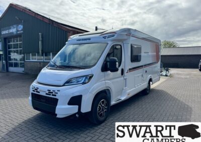 White camper van/motorhome parked on a brick lot beside a dark-metal workshop building with a 'Garage Swart' sign in the background.