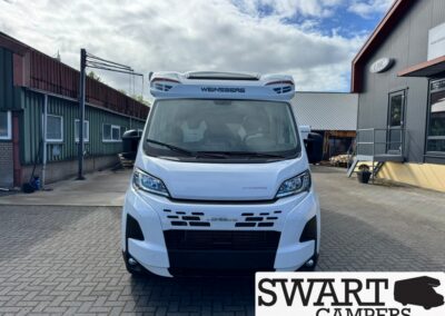Front view of a white camper van with a bold grille and modern headlights, parked in a paved lot beside industrial buildings; Swart Campers logo in the bottom right corner.