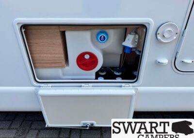 Open external service hatch on the side of a white camper van showing a water tank, red cap, blue inlet, and wood-panel interior