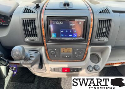 Dashboard of a camper van showing a center touchscreen infotainment unit, climate controls, and gear shifter with wood trim arc; SWART CAMPERS watermark.