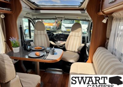 Cozy campervan interior with two front seats and a small table set for breakfast: two plates with croissants, cups, and a potted plant on a dark countertop, skylight above, SWART CAMPERS logo in corner.