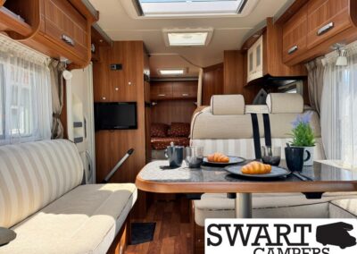Cozy camper van interior with beige striped seating around a wood dining table set for two, croissants and cups, and a skylight above.