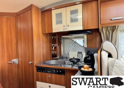 Compact camper van kitchen with wood cabinets, sink, two-burner stove, kettle, and a bowl of eggs on the counter near a rounded splash guard.