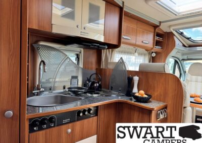 Interior of a wood-paneled camper van kitchen with sink, stove, kettle, and overhead storage cabinets