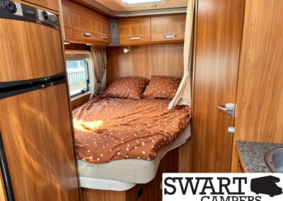 Wood-paneled camper bedroom with a full bed in brown polka-dot bedding, overhead cabinets, and a window with curtains; SWART CAMPERS watermark.