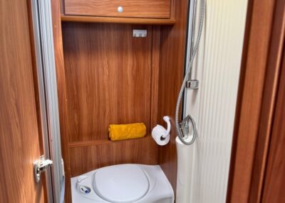 Compact camper bathroom with wood paneling, a small toilet, a handheld shower, a yellow towel on a shelf, and a cabinet above the stall door.