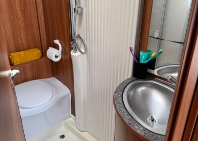 Compact camper bathroom with toilet, curved shower stall, and stainless sink set in a dark granite countertop; wood paneling surrounds.