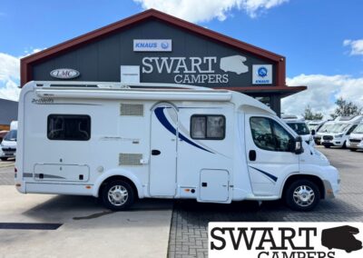 White motorhome parked in front of a Swart Campers building with blue decals and 'KNAUS' logos; dealership lot in background.