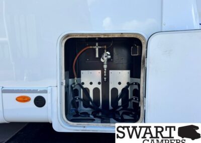 Open exterior storage bay of a white camper with hoses and metal fittings inside, visible valve assembly in the back wall.