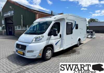 White motorhome with blue decals parked on a brick-paved lot beside a workshop under a blue sky.