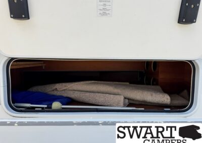 Open camper storage hatch revealing rolled carpet, blue fabric, and wood storage panels; SWART CAMPERS logo visible.