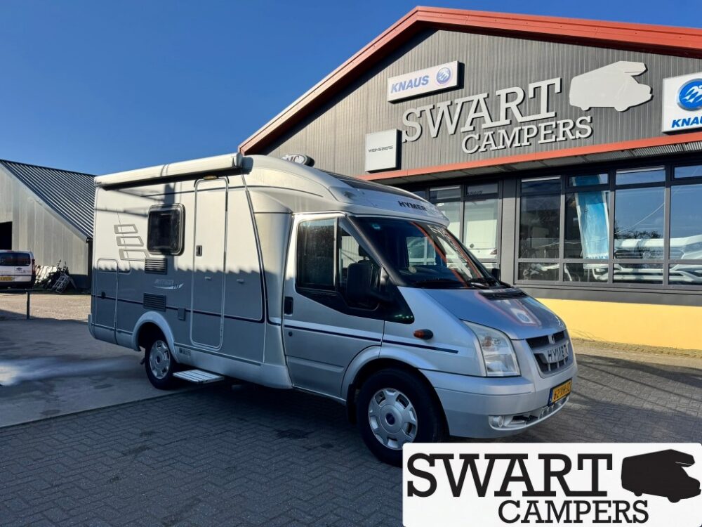 Silver motorhome parked outside a Swart Campers dealership building with logo and large windows in the background.