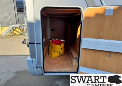 Open camper van door showing wooden floor storage with a yellow bag and a smiley on it.