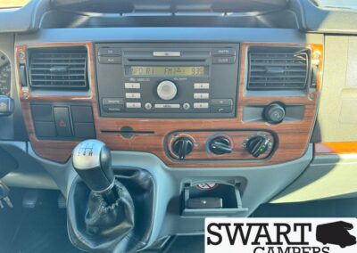 Dashboard of a vehicle with a manual gear shift, wood-toned trim, radio, and climate controls in the center.