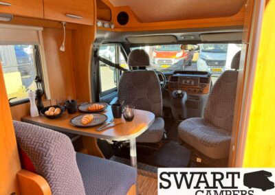 Interior of a camper van with a table set for breakfast: croissants, eggs, mugs, and fruit on plates near a cushioned bench and driver seats. (SWART CAMPERS watermark)