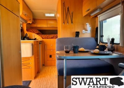 Interior of a wooden camper van with a dinette table set for breakfast—croissants, mugs, and a window view
