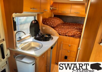 Wooden camper-van interior with a kitchenette (sink and two-burner stove), overhead cabinets, and a rear bed with brown polka-dot bedding.