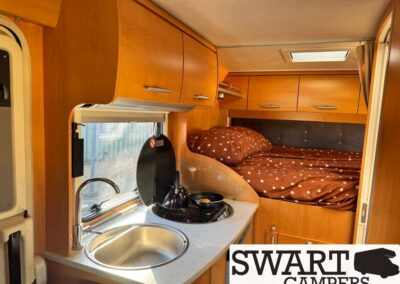 Cozy camper van interior with a compact kitchenette (sink, stovetop) and a raised bed with brown polka-dot bedding nearby.
