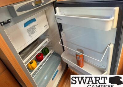 Open camper refrigerator showing peppers on the lower shelf and a can on the door shelf, with the freezer at the top.
