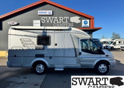 Silver motorhome parked outside a Swart Campers dealership with the SWART CAMPERS sign on the building edge and logo plaques nearby