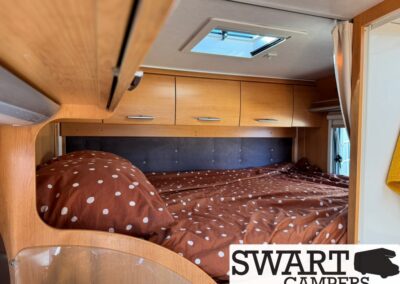 Camper van bedroom with brown polka-dot bedding, light wood overhead cabinets, and a skylight above the bed