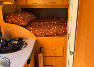 Compact camper interior with a wood kitchenette, brown polka-dot bedding, and upper storage cabinets above a small bed area