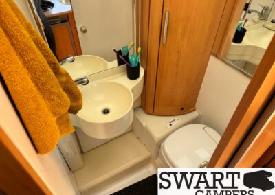 Compact camper bathroom with a round sink, orange towels, toothbrush holder, wooden cabinet, and a toilet corner.