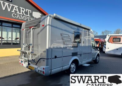 Grey motorhome parked outside a dealership with a rear bike rack and SWART CAMPERS logo visible on the building; other campers in the lot.
