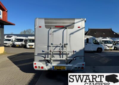 Rear view of a silver motorhome with a bike rack mounted on the back, in a camper lot with other motorhomes visible; clear blue sky above.