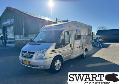 Silver Hymer motorhome parked in a dealership lot beside a workshop, with a SWART CAMPERS logo overlay in the corner.