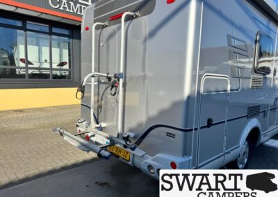 Grey motorhome with a rear-mounted bike rack, parked outside a camper dealership; Dutch license plate 02-XH-JZ.
