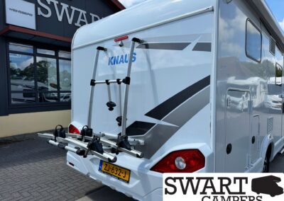 Rear view of a white KNAUS caravan with a bike rack mounted on the back, parked outside a dealership whose sign reads SWARTS CAMPERS (partial).