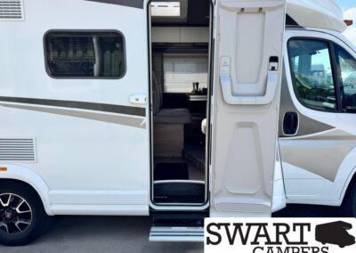 Open RV door reveals compact kitchenette and seating area inside a white motorhome. A Swart Campers sign is partially visible at the bottom right.