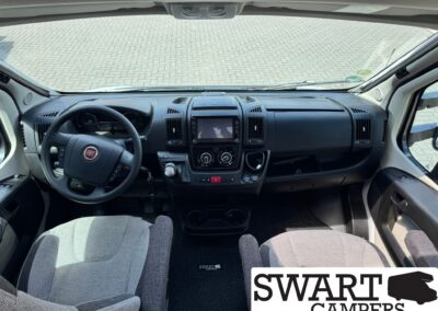 Front interior of a Fiat van: steering wheel on the left, dashboard, center touchscreen, and gray fabric seats with a floor mat reading SWART CAMPERS partially visible.
