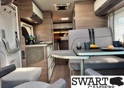 Cozy camper van interior with wood cabinets, a small kitchen, a table and gray seating, and a bed visible in the rear. A teddy bear sits on the bed.