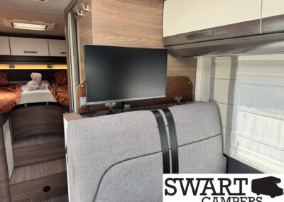 Interior of a camper van with a wall-mounted flat-screen TV, woodgrain cabinets, and a bed area with an orange duvet and a teddy bear on the bed.