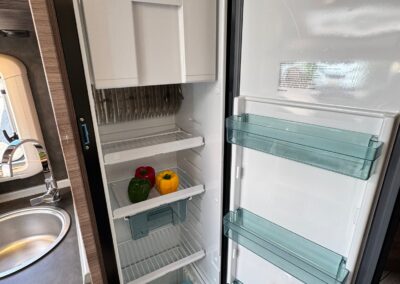 Open camper refrigerator with top freezer; inside are red, yellow, and green bell peppers on the lower shelf and an empty freezer drawer above.